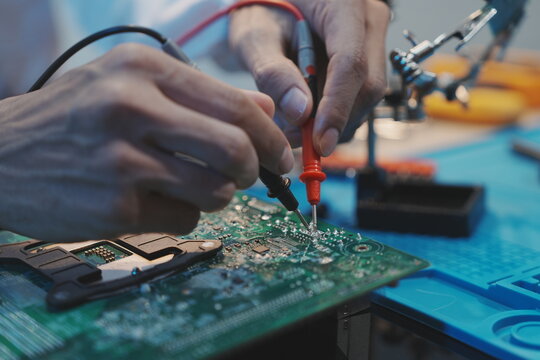 A close-up shot of a technician&rsquo;s hands using red and black multimeter probes to test specific electrical contact points on a green circuit board with precision.