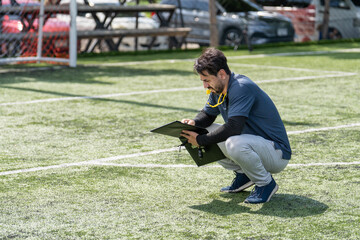 Fototapeta premium Professional soccer coach squatting on green turf field writing strategy on clipboard outdoor, football trainer or referee preparing tactical notes during match training session