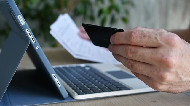 Person reads bank card agreement while holding credit card near tablet keyboard at home desk before online payment