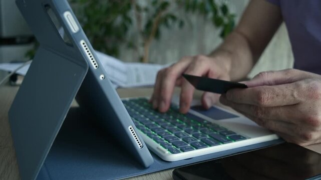 Person pays online at home with bank card, typing on tablet keyboard beside smartphone for secure digital checkout process