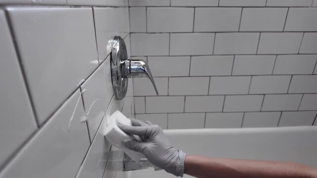 Close-up of a bathroom wall with white ceramic tiles being cleaned. A gloved hand uses a sponge to scrub the tile surface and grout lines. A bathtub and faucet are partially visible in the background