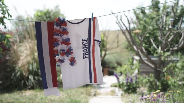 Soccer fan shirt, scarf and leis in national colors of France hanging outdoors in garden on clothesline on sunny windy day
