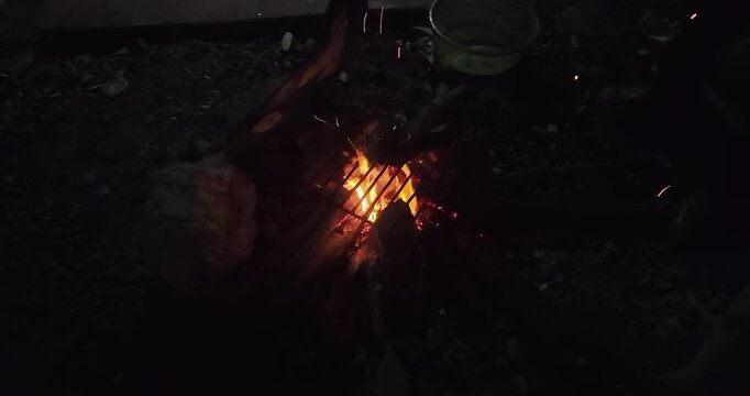 Close up: a person turns two fish grilling over crackling flames and flying sparks on a small charcoal grill at night.