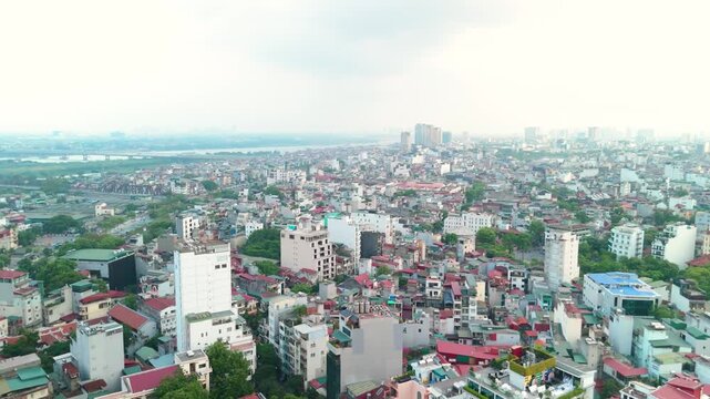 Hanoi city expanding along the red river shoreline, revealing urban density, residential rooftops, modern buildings, and the distant long bien bridge spanning the wide waterway
