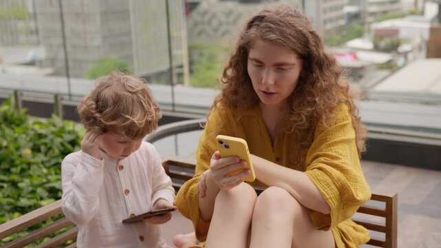 Freelance mother on a video conference call from an outdoor hotel balcony overlooking a city with her son playing on his smartphone