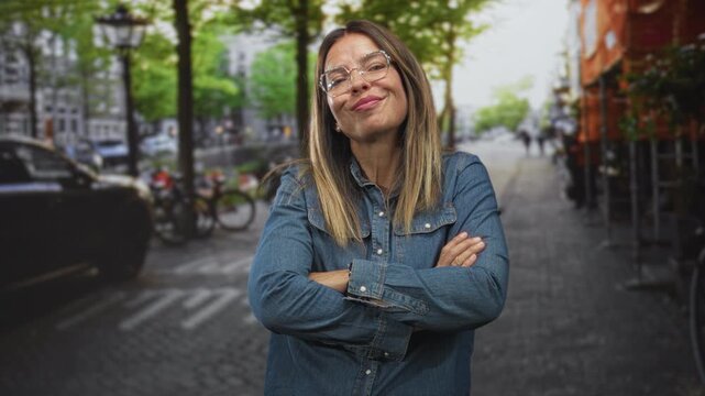 Woman wearing glasses with arms crossed and slight smirk on a cobblestone street near parked bicycles and a sidewalk cafe; confidence attitude.