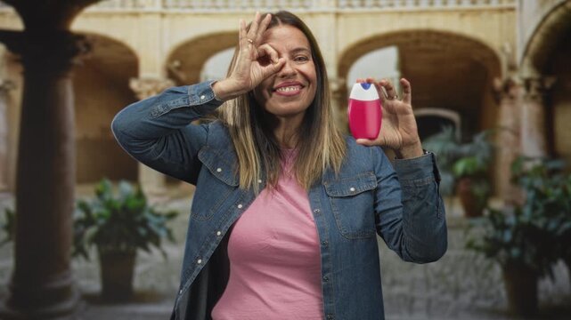 Woman holding a toothpaste bottle and making an ok sign over her eye in a building courtyard; playful confidence.