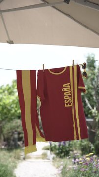 Soccer fan shirt, scarf and sunglasses in red and yellow national colors of Spain hanging on line under parasol outdoors in garden on sunny day for drying, background blurred
