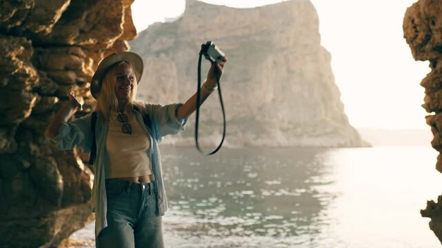 Happy female traveler filming herself with retro camera inside sea cave during sunset, creating travel content and sharing authentic vacation experiences.