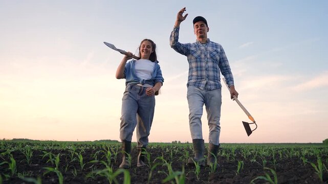 A man and a woman walk together in a green field, both smiling and engaging with each other. The man holds a tool while the woman carries a shovel, both enjoying the agricultural setting.