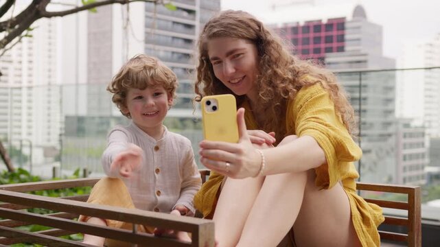Curly young mother and son wave hello and goodbye during video call from hotel balcony in big city