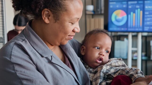 African American woman holding baby while reviewing documents in office with charts on screen. Ideal for campaigns on remote work, working mothers, and hybrid employment