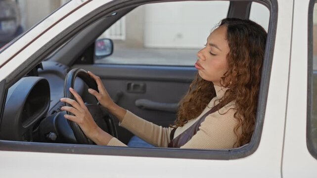 Young latina woman leaning head on steering wheel, hand gripping wheel, slumped in parked white car on street; fatigue resignation.
