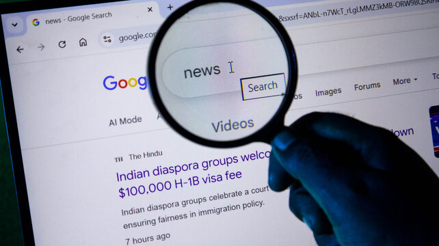 Closeup of a hand holding a magnifying glass over a computer screen showing a google search for news, with a headline about h1b visa fees