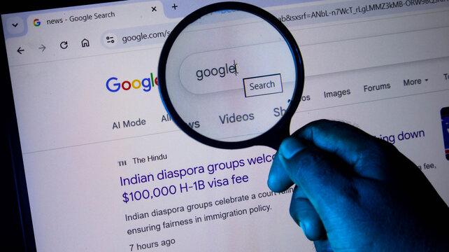 Closeup of a hand holding a magnifying glass over a computer screen showing a google search page with a news headline about h1b visa fees