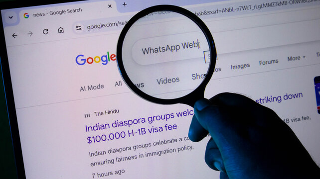 Closeup of a hand holding a magnifying glass over a computer screen showing a google search page for whatsapp web and news about h1b visa fees