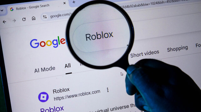 A person holding a magnifying glass over a computer screen displaying a google search for roblox, focusing on the search term