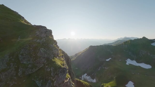FPV drone flight moving forward along a steep alpine cliff at sunrise, revealing green ridges, snow patches, distant valleys and warm morning light under a clear mountain sky