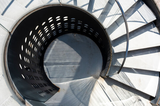 Circular staircase descends inside a spiral form, with metal railing, steps, shadow, light, pattern, architecture and geometric depth