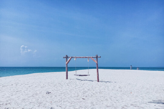 A wooden swing set stands on a pristine white sand beach under a clear blue sky