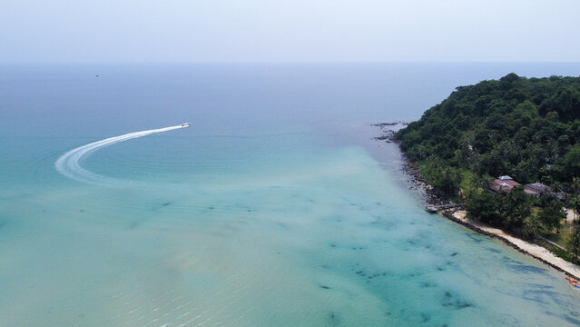 An aerial drone view of a speedboat leaving a white wake curve in the turquoise sea next to a lush green tropical island.
