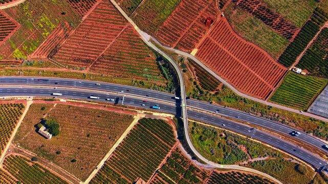 Aerial perspective of curved bridge crossing highway in agricultural region. High angle drone view of concrete overpass structure over multi lane motorway surrounded by patterned red soil fields