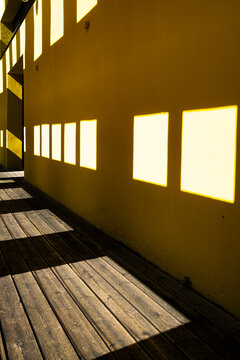 Window shadows fall across a wooden walkway beside a yellow wall, with railing, facade, courtyard architecture and geometric light