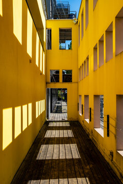 A narrow walkway runs through a yellow courtyard facade, where windows, balconies, shadows and sunlight form urban geometry along the wall