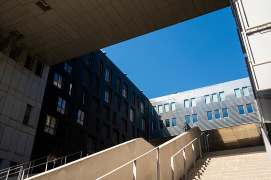 Blue sky cuts between urban buildings above concrete stairs and ramp, with railing, windows, shadow and courtyard architecture in a tight passage