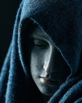 Extreme close up profile of a weeping Virgin Mary statue carved from grey stone, wearing a rustic blue burlap hooded cloak in dramatic moody light