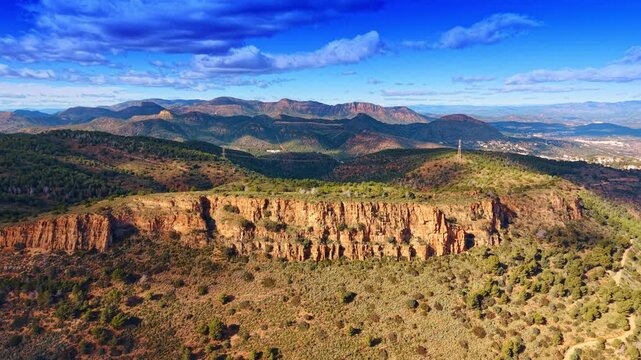 Scenic landscape of rocky cliffs and mountainous terrain under clouds. Wide angle view of orange limestone cliffs, scrubby vegetation, and distant mountain ridges under blue sky.