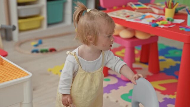 Girl holding orange stool beside red play table and foam alphabet mat in building; playful curiosity.