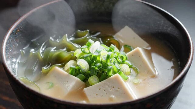 Steaming hot miso soup with tofu cubes, wakame seaweed, and fresh green onions in a dark bowl
