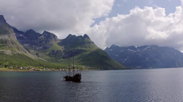 A vintage sailing ship navigates a peaceful fjord, framed by majestic green mountains and a dramatic cloudy sky, evoking a sense of timeless adventure and natural splendor