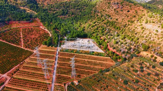Electrical substation and transmission towers in an orange orchard. High angle detail of power grid infrastructure surrounded by structured rows of citrus trees and red soil.