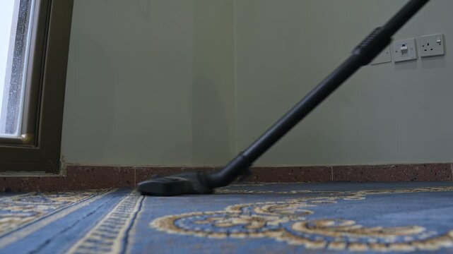 Low angle close up of a vacuum cleaner wand cleaning a blue patterned carpet next to a wall. Daylight shines from a nearby window during indoor floor maintenance and routine housekeeping.