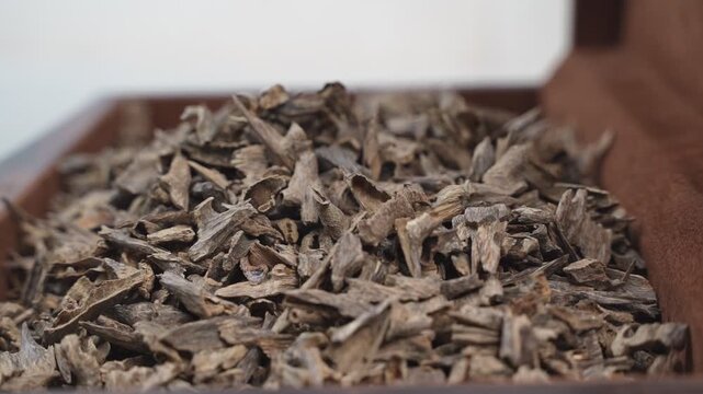 Close up cinematic shot of a person's hand sifting through a large pile of dry agarwood oud chips inside a storage box. The shallow depth of field highlights the texture of the aromatic incense.