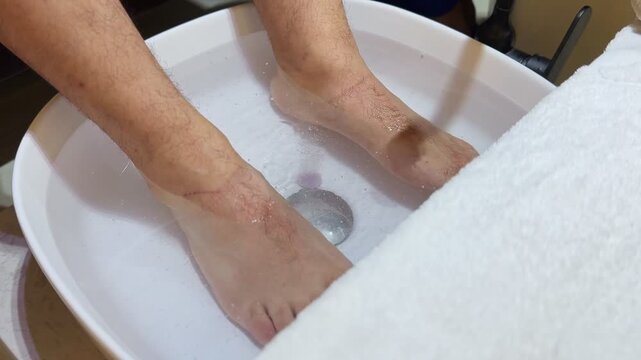 a man soaking his bare hairy feet in a white ceramic pedicure basin filled with warm water. Preparing for a relaxing spa treatment indoors with a fresh white towel resting nearby.
