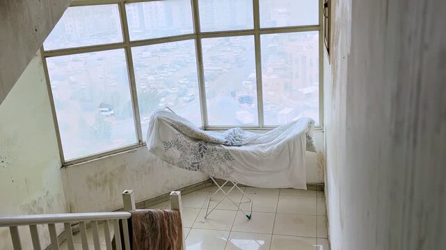 Wide shot of a floral blanket drying on a metal rack by a large window in a run-down indoor stairwell. Daylight reveals peeling paint walls and a hazy view of a busy city parking lot outside.
