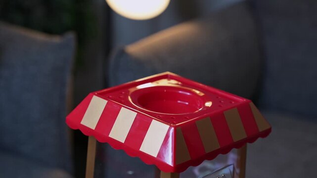 Close-up of a hand removing the red lid from a miniature striped popcorn machine. The retro carnival toy sits indoors against a blurred dark living room background with warm artificial lamp light.