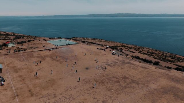 Drone view of young people playing soccer on a hillside field on Isla del Sol, with Lake Titicaca and the Bolivian Andes in the background.