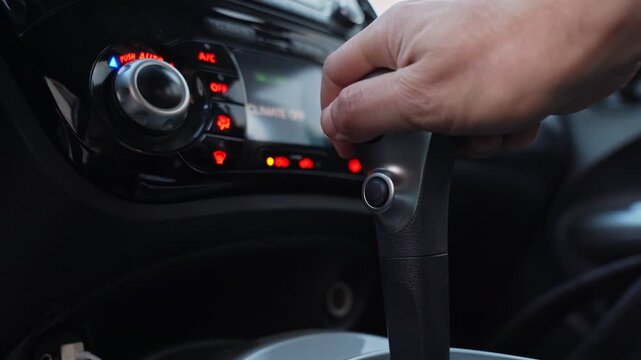 Close up shot of a driver's hand shifting the automatic gear selector inside a modern vehicle. The dashboard climate control illuminates the blurred background with a shallow depth of field.