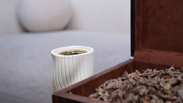 Close up of a modern white ceramic mabkhara beside a velvet-lined box filled with oud chips. Soft indoor daylight illuminates this traditional home fragrance setup against a blurred bright background