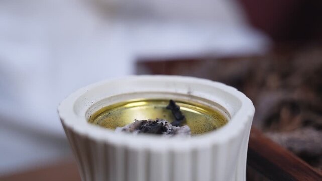 Extreme close-up of burnt bakhoor and ash resting on used charcoal inside a white ceramic incense burner. Soft studio lighting highlights the gold interior against a blurred background of oud chips.