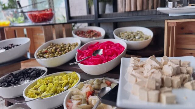 a colorful Mediterranean restaurant buffet featuring white bowls filled with black and green olives, pink pickled turnips, yellow peppers, and cheese cubes on a dark wooden table indoors.