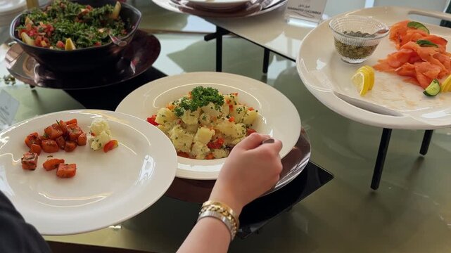 a person's hand wearing bracelets using a spoon to serve fresh potato salad onto a white plate. The bright daytime hotel buffet also features smoked salmon and healthy green appetizers.