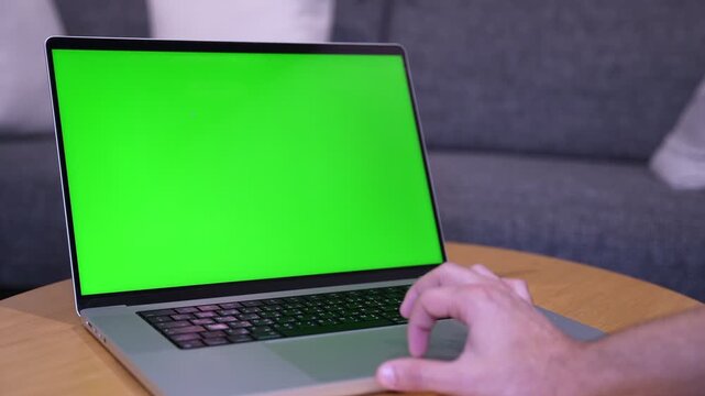 Close-up of a hand using the trackpad of a green screen laptop resting on a wooden table. The modern computer sits in an indoor living room with a blurred grey sofa in the background.