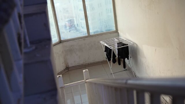 High-angle shot looking past a blurred railing at a folding laundry rack with clothes drying. The rack sits on a tile floor in a dirty, sunlit apartment stairwell with large windows during daylight.