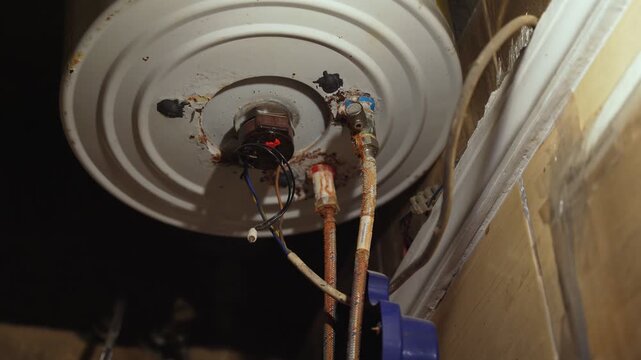 Close up of old electric water heater underside showing rusted pipes, exposed electrical wiring, and corrosion buildup. Indoor plumbing and heating system in poor maintenance condition.