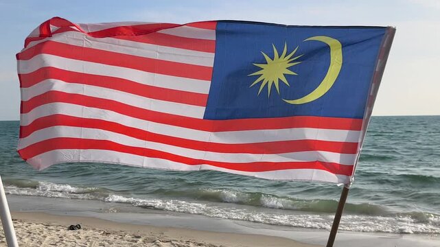 The Malaysian flag stands proudly on a sandy beach. Strong coastal winds blow the flag fiercely against the sky. This powerful display symbolizes national pride and freedom by the sea.